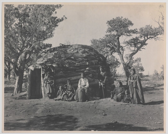A black-and-white photograph of a group of Indigenous adults and children in front of a round, wood dwelling.