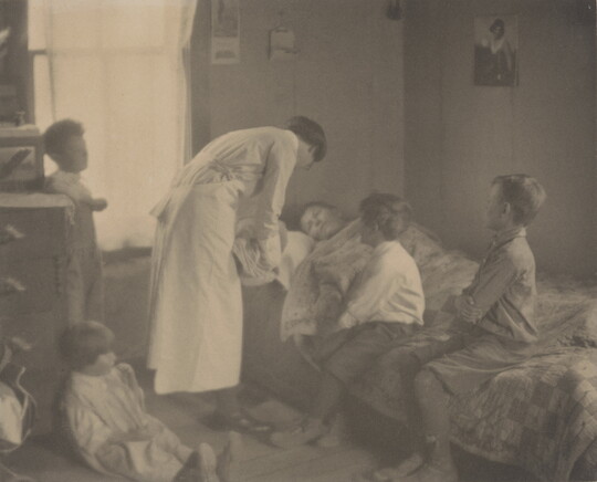 A black-and-white photograph of a nurse bending over a person in a bed, two children sitting on the bed, and two children behind the nurse.