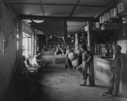 A black-and-white photograph of Indigenous people in a shop with a variety of products such as animal hides, tin pails, and lanterns hanging from the ceiling.