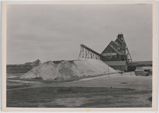 A black-and-white photograph of large pile of salt in front of a multi-story industrial building with a sign that reads "Crystal Salt Co."