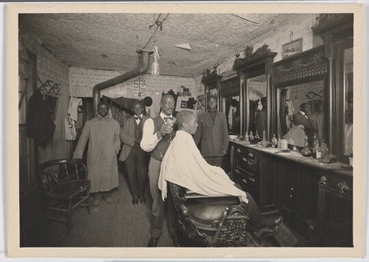 A black-and-white snapshot of the inside of a barber shop with a Black man in a chair getting his hair cut.