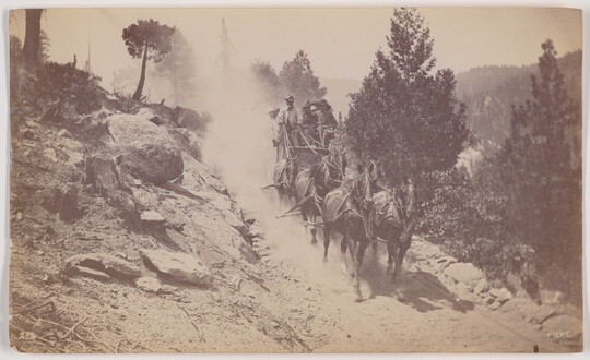 A black-and-white photograph of a stagecoach being pulled by six horses down a dusty dirt road on a hillside.