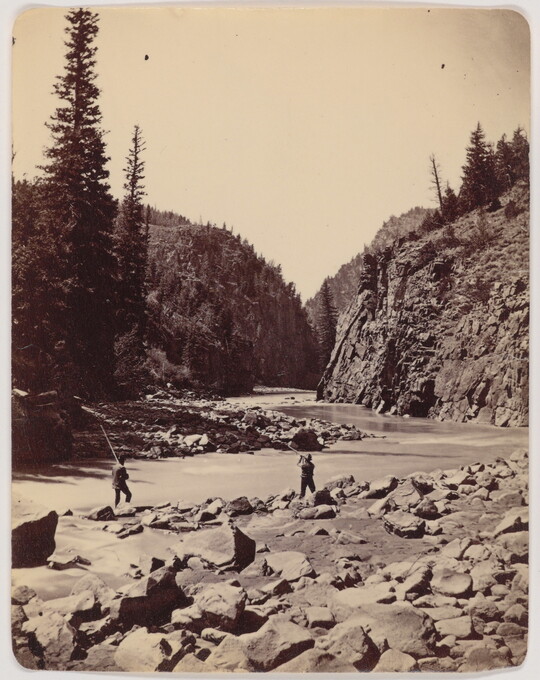 A black-and-white photograph of two men fishing in a river surrounded by a rocky shore and cliffs.