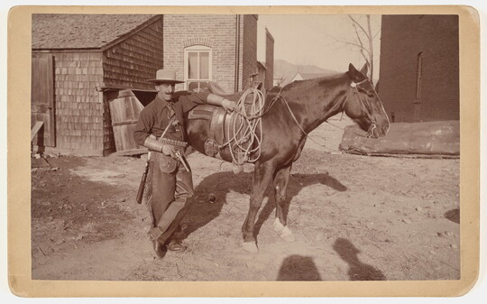 A black-and-white photograph of a light-skinned man with a large mustache, wearing a cowboy hat, holding a gun and leaning against a horse.
