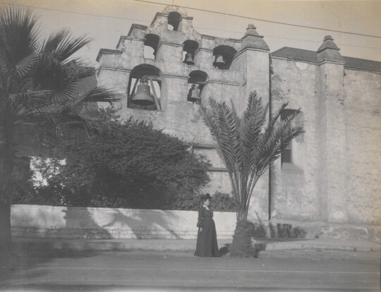 A black-and-white photograph of a woman standing next to a building with several large bells hanging near the top.
