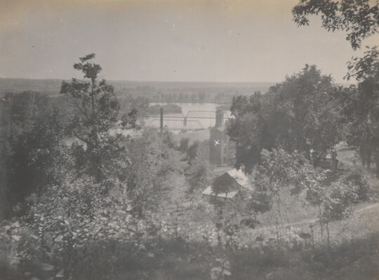 A black-and-white photograph taken from a higher elevation of a bridge spanning a river and forested area that includes some buildings.