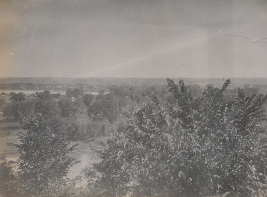 A black-and-white photograph taken from a higher elevation of a forested area and a river in the distance.