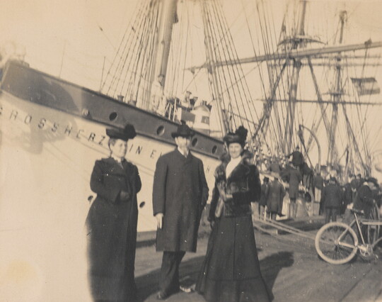 A black-and-white photograph of three nicely dressed adults standing on a dock in front of a massive ship.