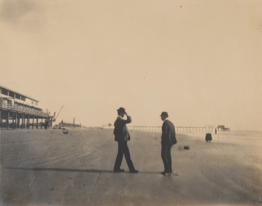 A black-and-white photograph of two men in bowler hats and suits standing on a sandy beach.