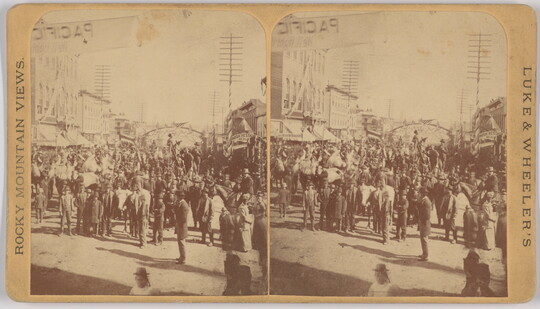 A sepia-toned stereograph of a large gathering of people in a street lined with buildings.