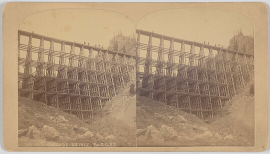 A sepia-toned stereograph of a high trestle bridge over a mountainous ravine viewed from the bottom.