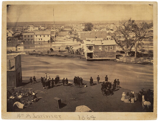 A sepia-toned photograph of damaged buildings and streets surrounded by mud and water as people look at the devastation.
