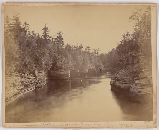 A black-and-white photograph of a wide calm river lined with trees and rock formations.