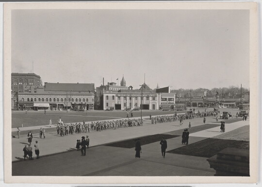 A black-and-white photograph of a parade coming through a wide street as scattered spectators watch.