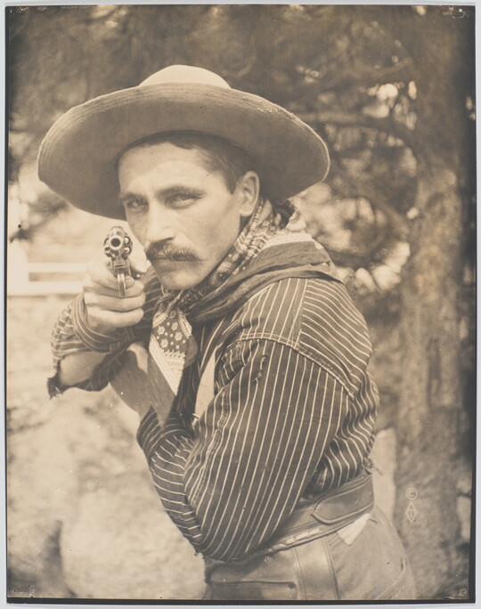 A sepia-toned photograph of a White man in a cowboy hat and bandana pointing a revolver at the camera.