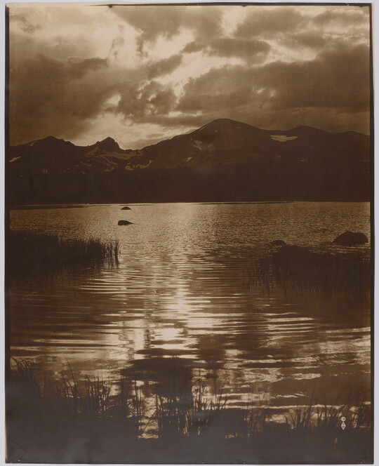 A sepia-toned photograph of light filtered through clouds reflecting on a lake surrounded by mountains.