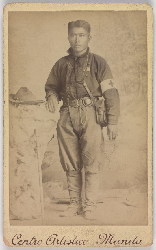 A black-and-white photograph of a medium-skinned soldier with a canteen slung across his chest and wearing a Red Cross armband.