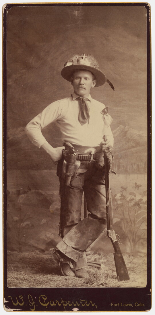 A sepia-toned studio portrait photograph of a cowboy wearing a hat decorated with feathers, a belt holding a revolver, and leaning on the muzzle of a rifle.