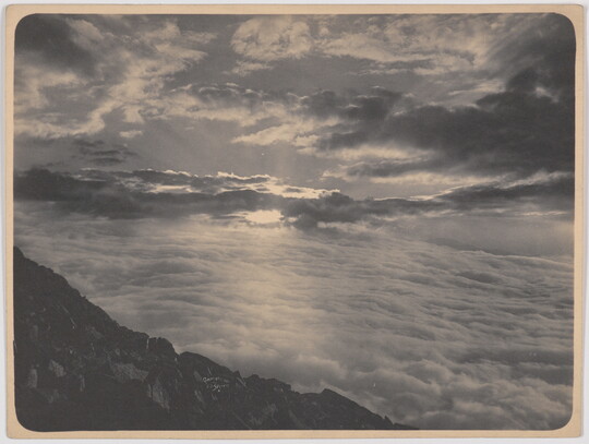 A black-and-white photograph of a dramatic cloudscape with the sun and rays peeking through below a rocky landscape in the foreground.