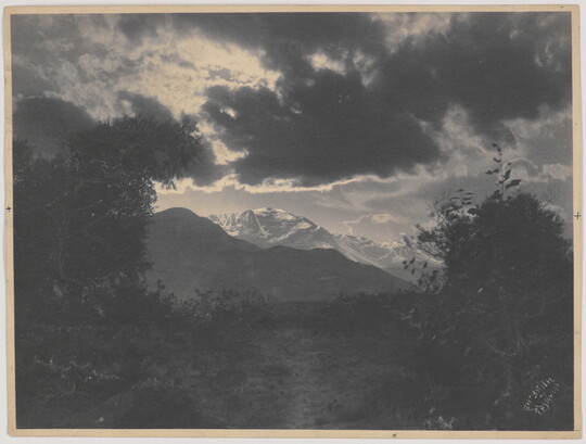 A black-and-white photograph of snow-capped mountains under dark and sun-lit clouds framed by trees.