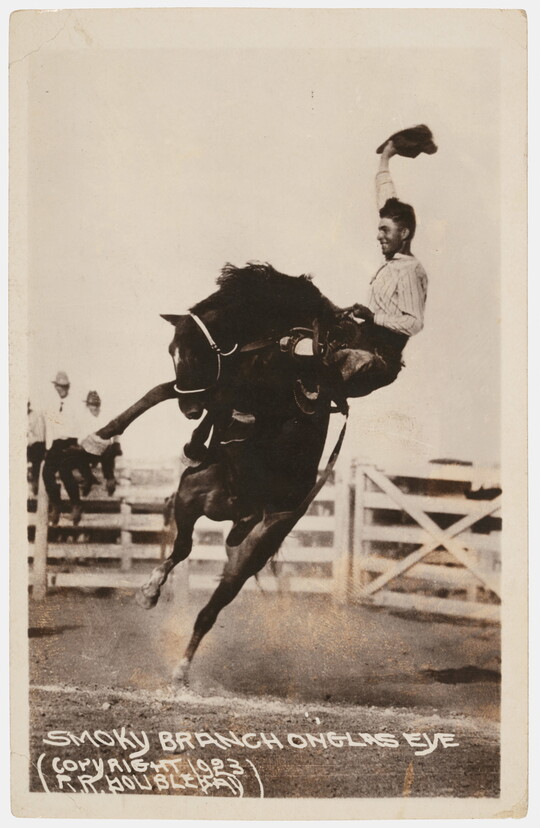 A black-and-white photograph of a cowboy waving his hat as he holds on to a rearing horse at an outdoor rodeo.