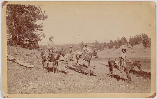 A sepia-toned photograph of several cowboys on horseback hauling large pieces of wood with ropes.