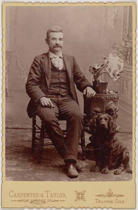 A black-and-white portrait photograph of a seated White man with a mustache wearing a three-piece suit and a dog sitting at his feet.