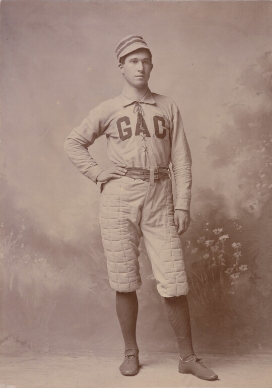 A sepia-toned portrait photograph of a White man, one hand on hip, wearing a baseball hat and uniform with "GAC" across the chest.