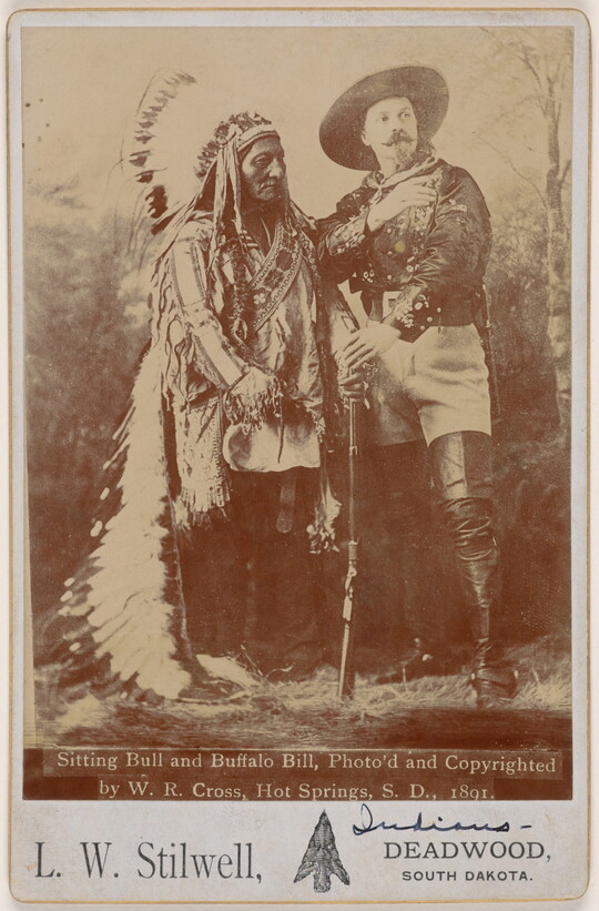 A sepia-toned portrait photograph of an Indigenous man wearing a feathered headdress and traditional clothing next to a White man in a large hat and tall leather boots holding a rifle in front of them.