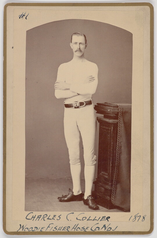 A black-and-white portrait photograph of a young White man with a mustache wearing all white standing with arms crossed next to a decorative column.