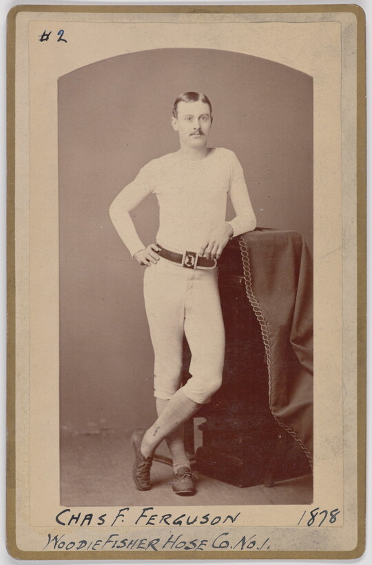 A black-and-white portrait photograph of a young White man with a mustache wearing all white, leaning casually against a decorative column.