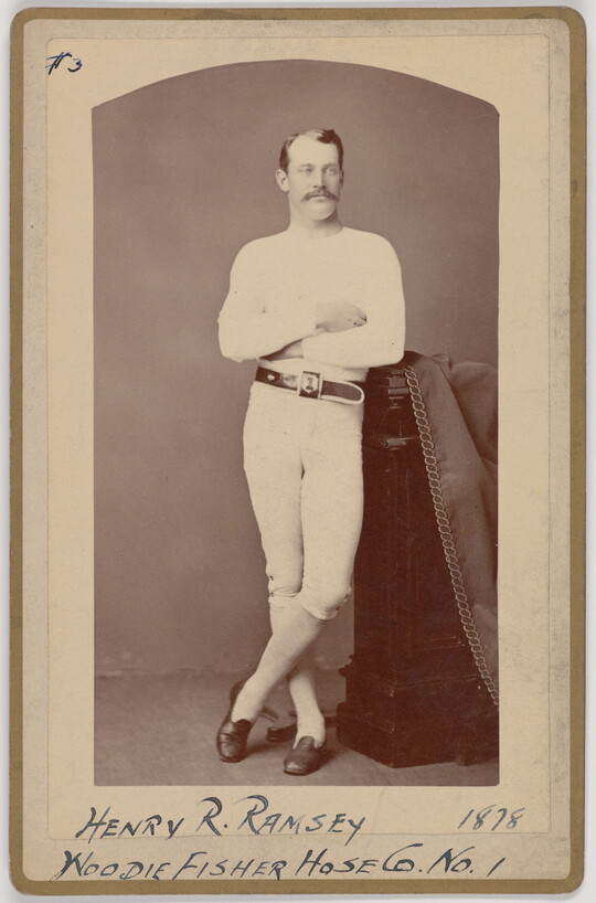 A black-and-white portrait photograph of a young White man with a mustache wearing all white standing with arms crossed leaning his hip on a decorative column.