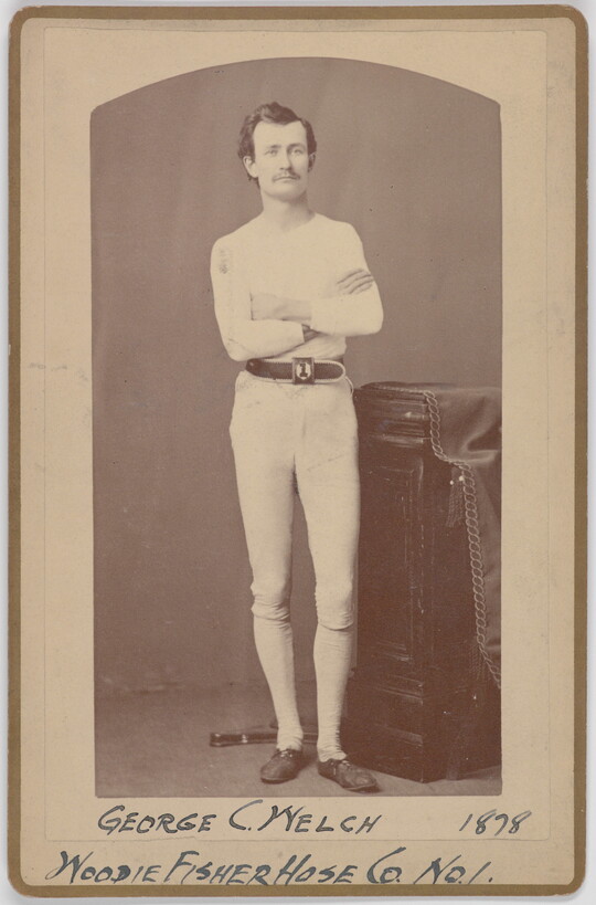 A black-and-white portrait photograph of a young White man with a mustache wearing all white standing with arms crossed next to a decorative column.