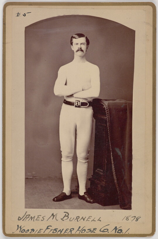 A black-and-white portrait photograph of a young White man with a mustache wearing all white standing with arms crossed next to a decorative column.