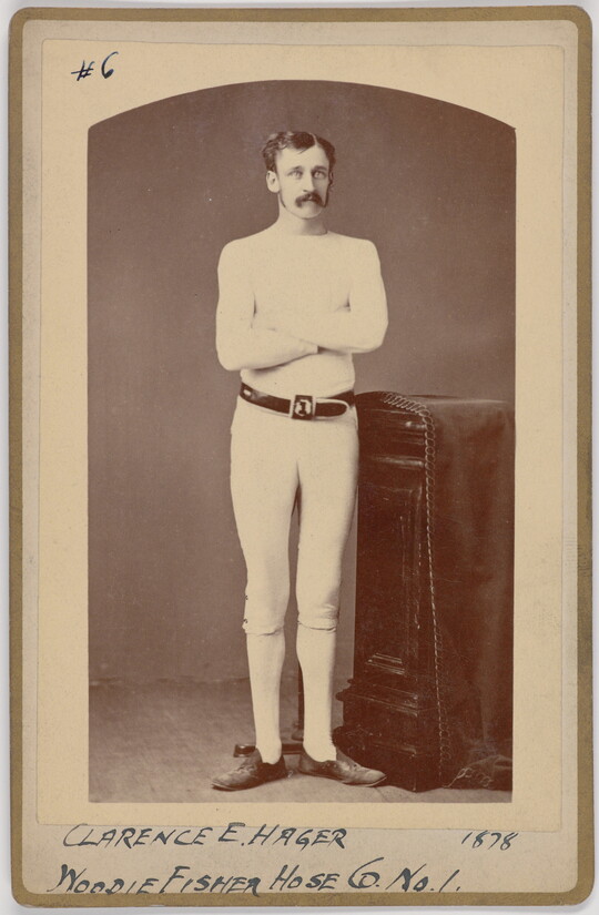 A black-and-white portrait photograph of a young White man with a mustache wearing all white standing with arms crossed next to a decorative column.