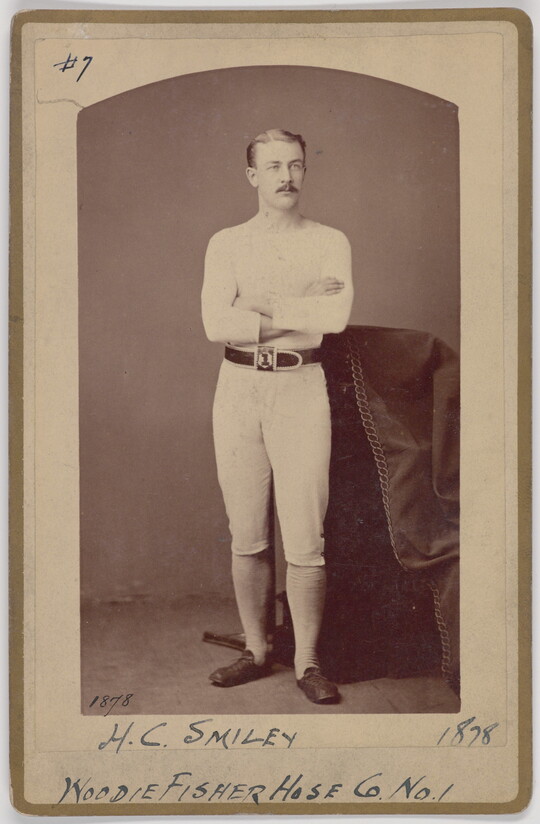A black-and-white portrait photograph of a young White man with a mustache wearing all white standing with arms crossed next to a decorative column.