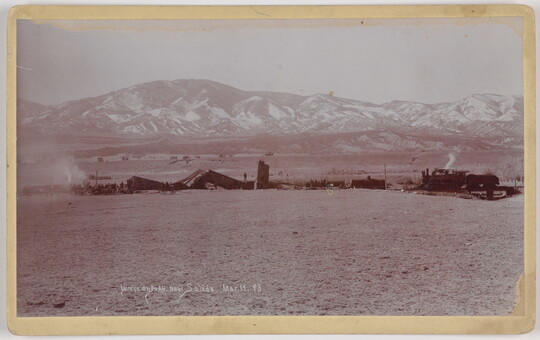 A black-and-white photograph of mangled train cars and an engine on a plain with snow-covered mountains in the distance.
