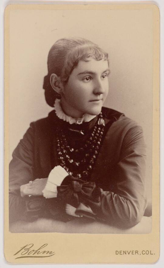 A black-and-white portrait photograph of a young White woman, hair pulled back and short bangs framing her face, wearing a dark dress with a ruffled white collar and sleeves, leaning with her elbows on the surface in front of her.