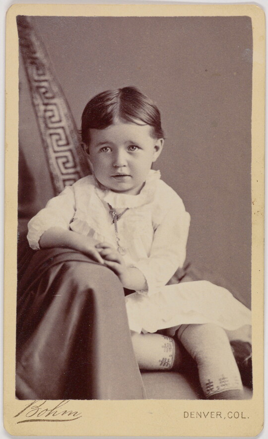 A black-and-white portrait photograph of a formally-dressed White child posed on a chair draped in fabric.
