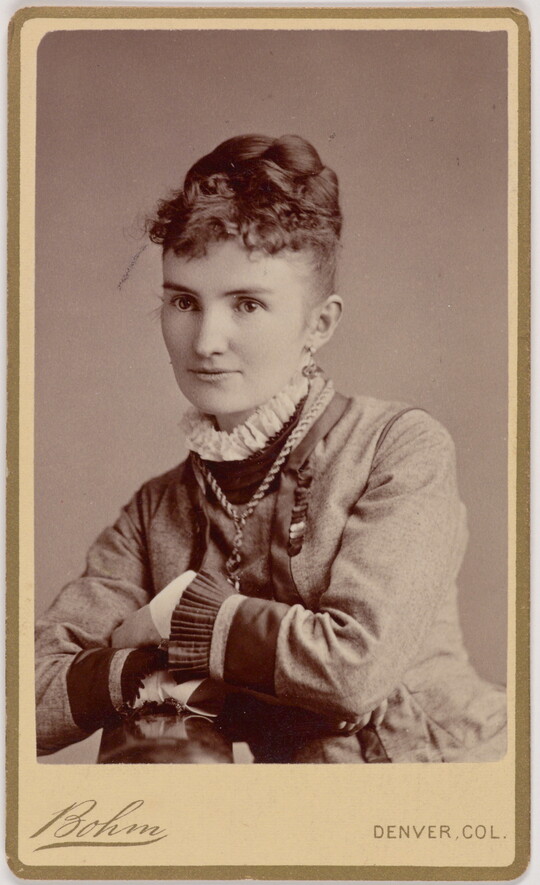 A black-and-white portrait photograph of a young White woman, hair piled on top of her head, wearing a dark dress with a ruffled white collar and sleeves, leaning with her elbows on the surface in front of her.