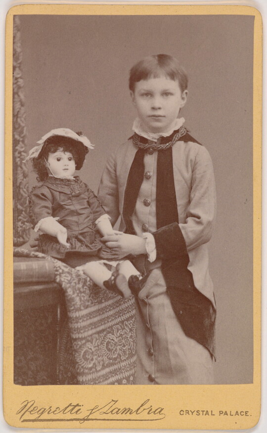 A black-and-white portrait of a young White child with short hair standing next to a seated doll.