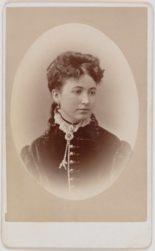 A black-and-white oval-shaped portrait photograph of a White woman, hair pulled up with ringlets that lay on her shoulders, wearing a dress with a high ruffled neck.