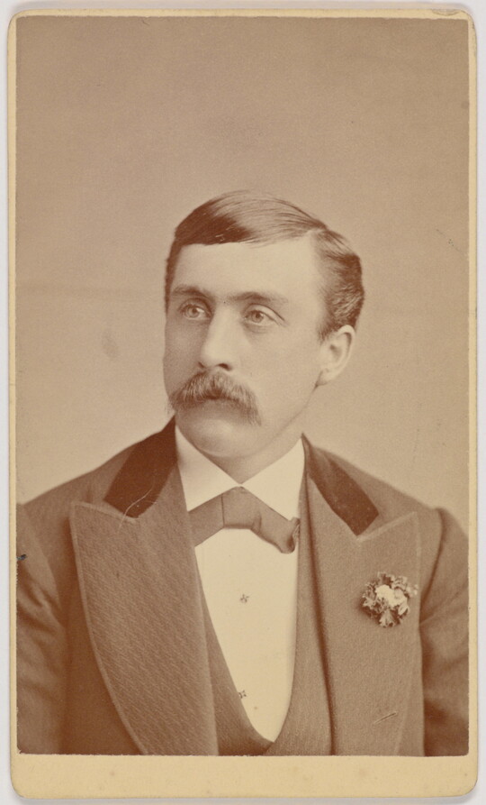 A black-and-white portrait photograph of a formally dressed White man with a mustache, wearing a corsage pinned to his lapel.
