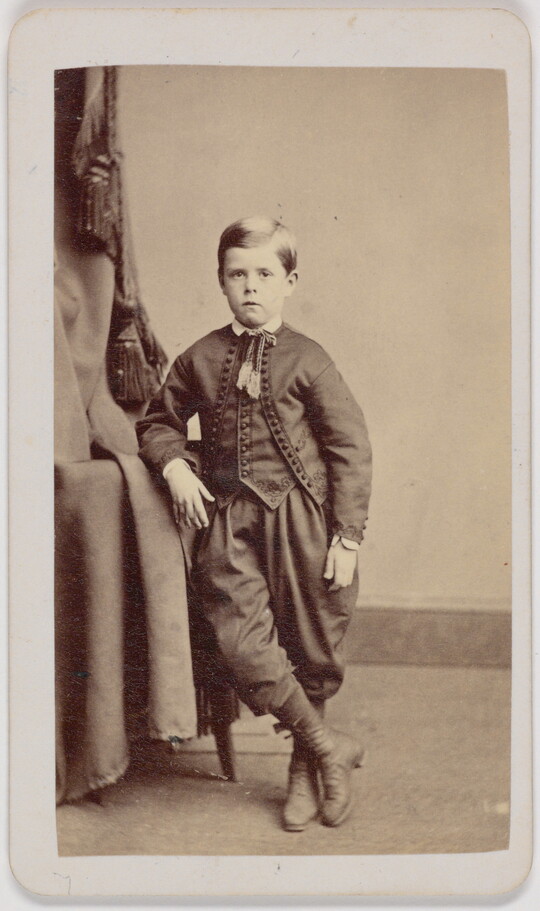 A black-and-white portrait photograph of a White boy wearing a formal jacket and vest over knickerbocker pants, leaning on a table, one leg crossed over the other.