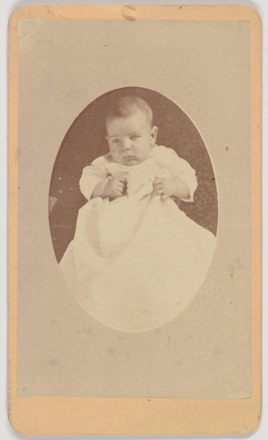 A black-and-white oval-shaped portrait photograph of a White infant in a white gown.