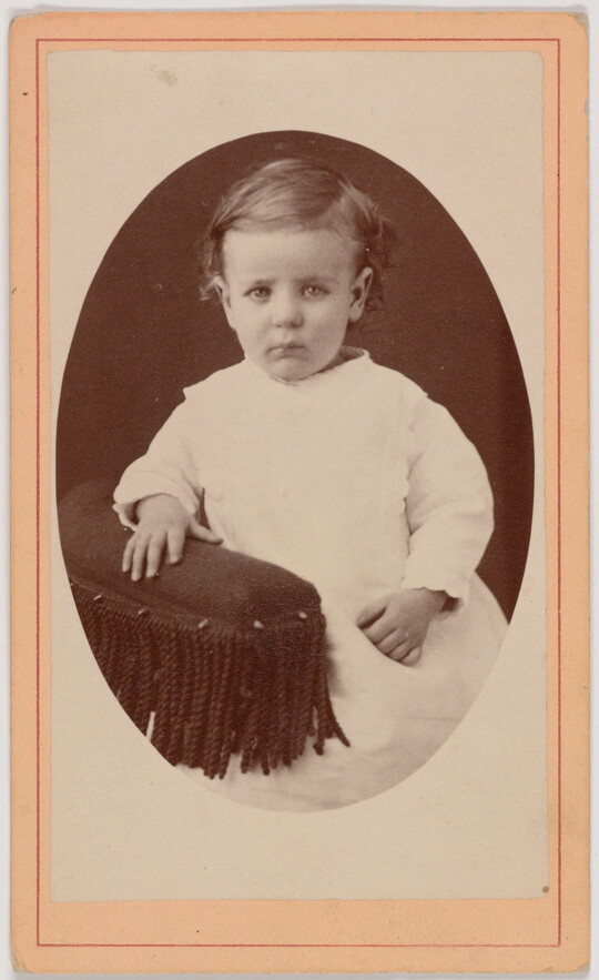 A black-and-white oval-shaped portrait photograph of a White toddler in a white dress resting one hand on the fringed arm of a chair.