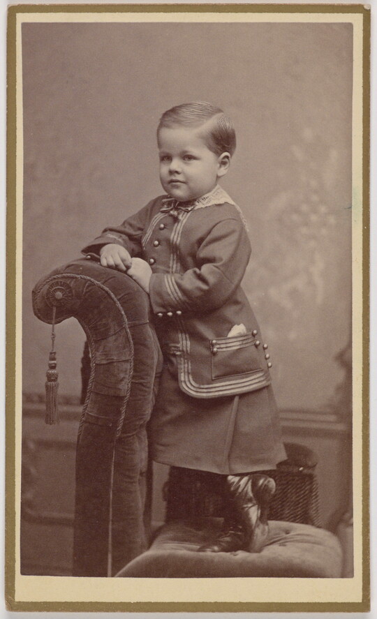 A black-and-white portrait photograph of a very young White child wearing what looks like a jumper under a formal jacket, standing on an upholstered chair and leaning on its back.