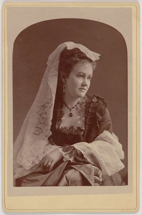 A black-and-white portrait photograph of a young White woman with a white lace veil on her head, wearing a dress with a square neckline trimmed in lace, her arms crossed on her lap.