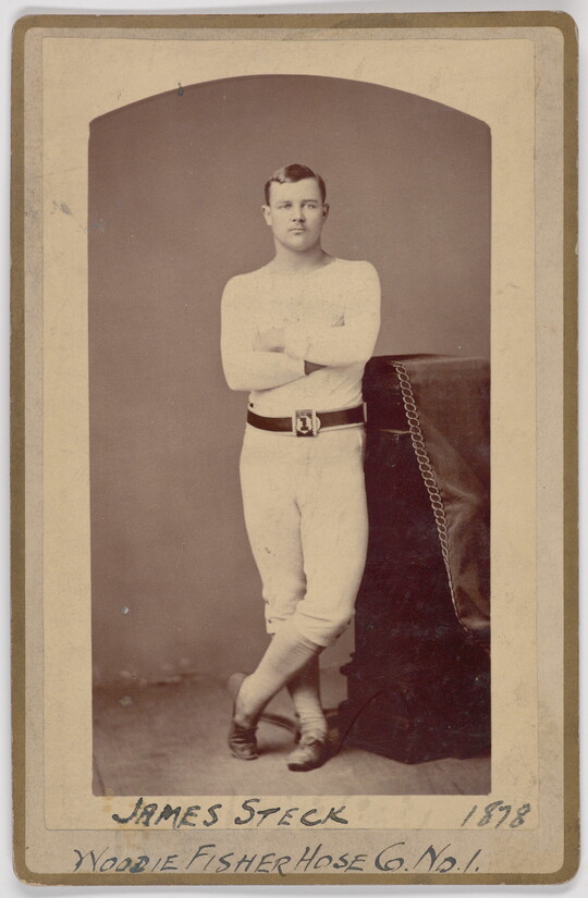 A black-and-white portrait photograph of a young White man wearing all white standing with arms crossed leaning his hip on a decorative column.