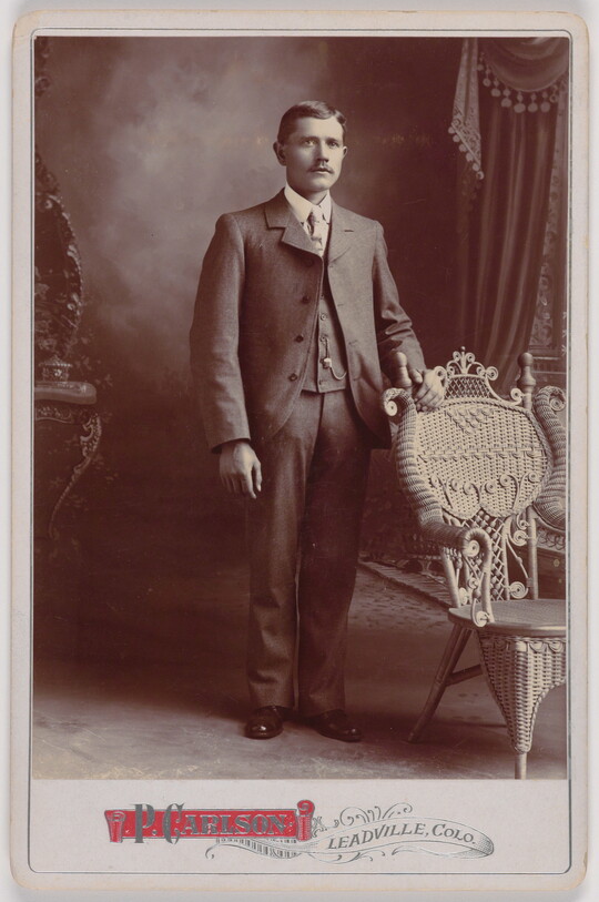 A black-and-white portrait photograph of a White man wearing a three-piece suit standing next to a wicker chair on which he rests his hand.